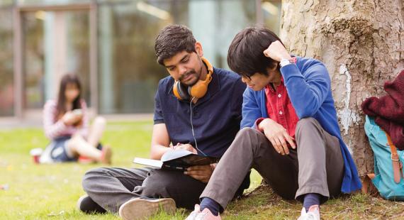 Two students at the University of Salford, sat under a tree and looking at a book together.