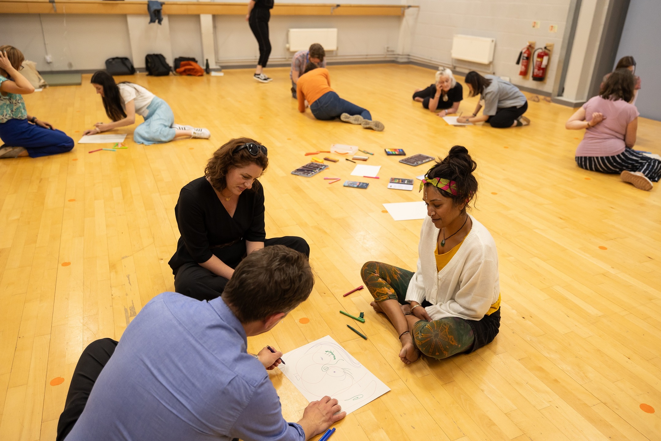 Groups of women are sat on the floor in a hall writing on paper, taking part in a workshop