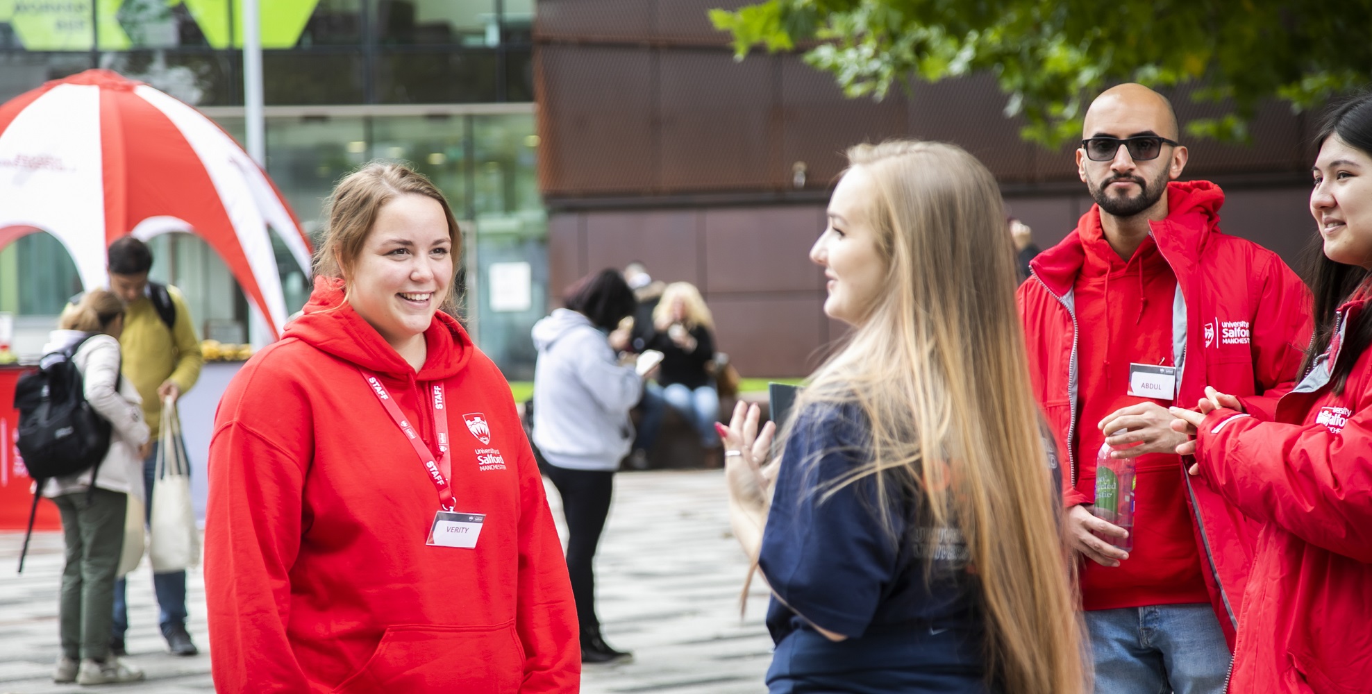 Three student ambassadors, in red University-branded hoodies, chatting to member of staff with concrete University building in the background