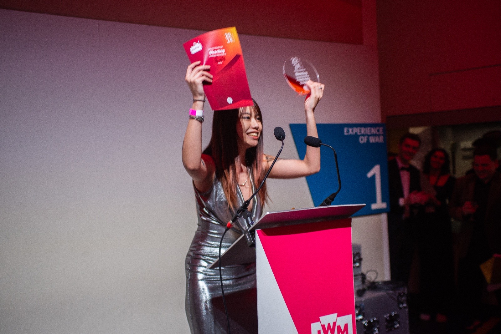 A woman in a silver dress holds a piece of paper and a trophy at an awards ceremony