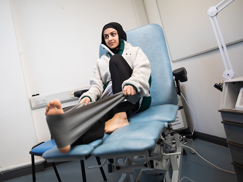 Seated on a clinical chair, a student demonstrates an exercise by stretching her leg and foot with a large exercise band.
