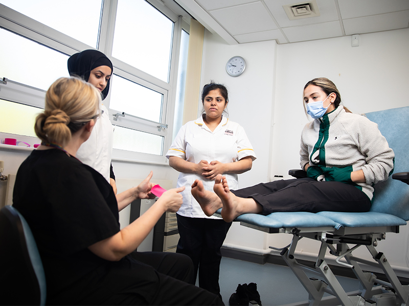 A tutor demonstrates a podiatry procedure while one student sits on a clinical chair, observed by two others.