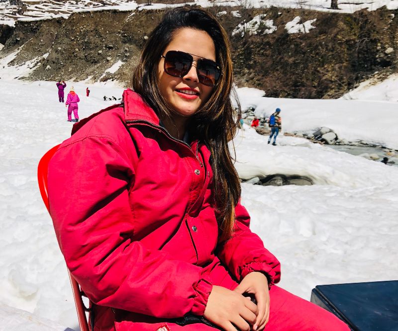 A female student in a ski resort wearing a pink ski suit and brown oversized sunglasses smiling at the camera.