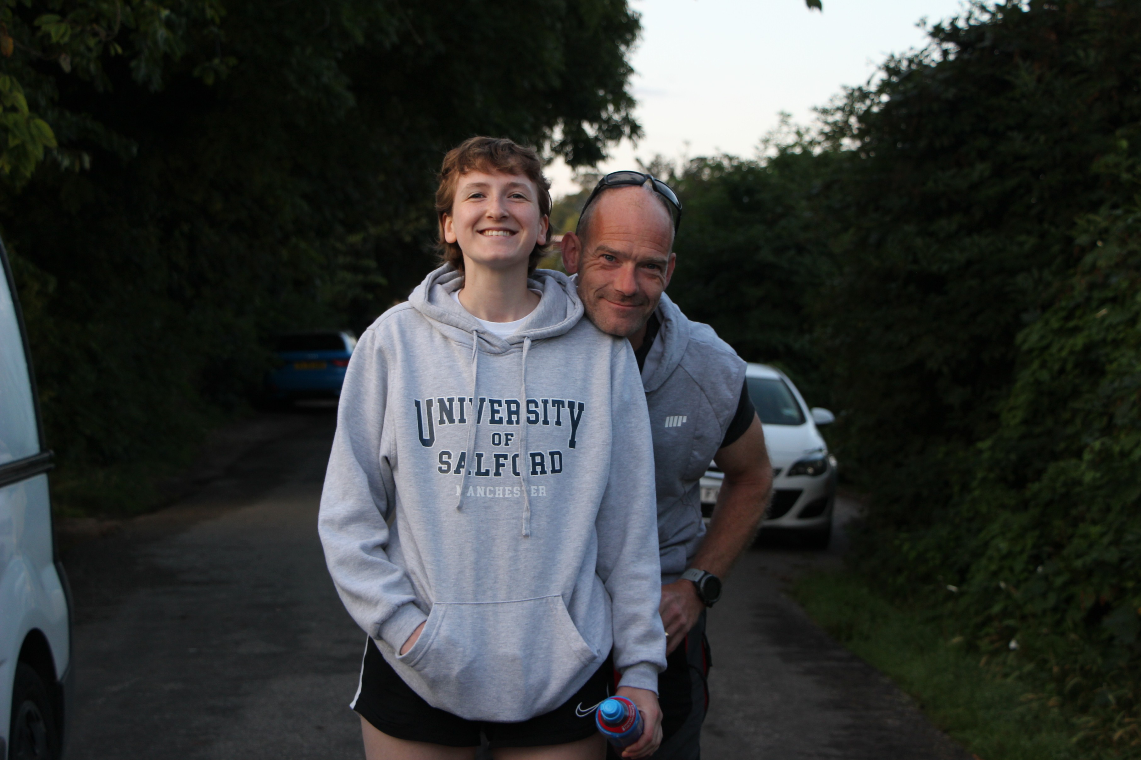 A girl in a grey jumper stands on a road with her father in the background