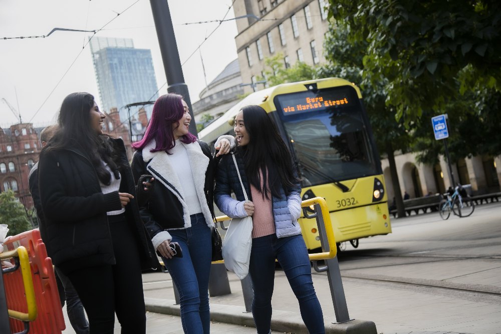 Three students walking around Manchester city centre alongside a tram