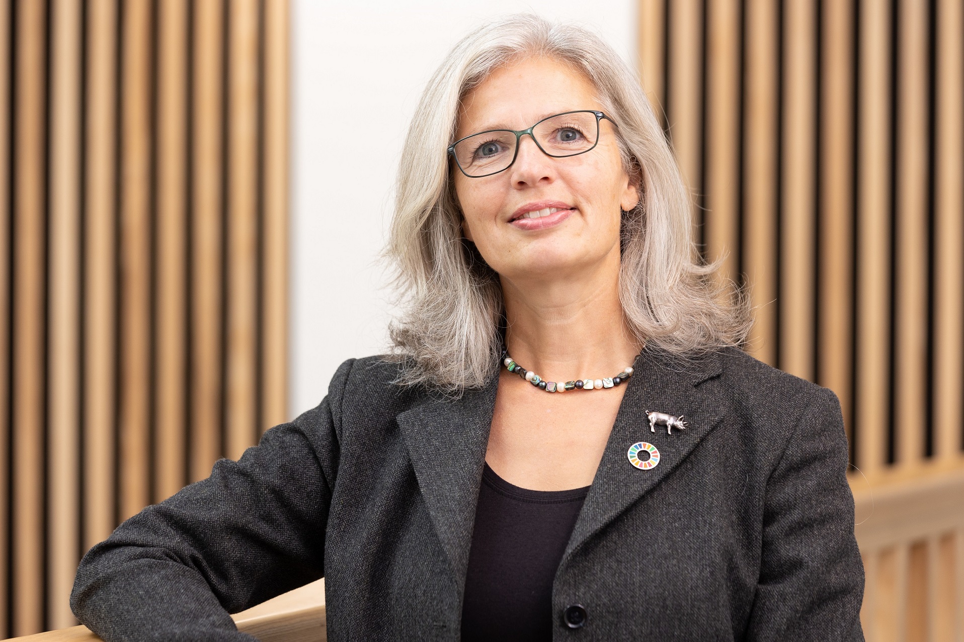 Professor Katy Mason smiling into the camera, wearing a blazer and stood in a room with acoustic paneled walls