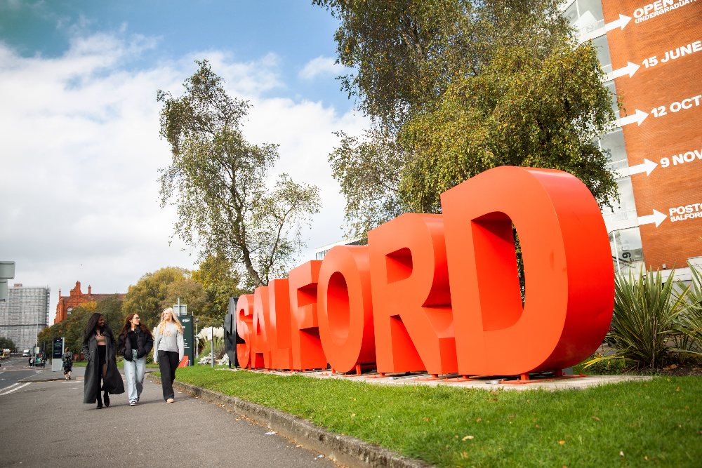 Three female students walking down the street next to a sign which says Salford in big red letters