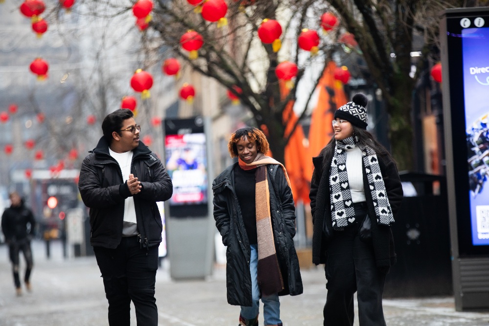 A male student chatting with two female students walking down a street with red lanterns on the trees behind them