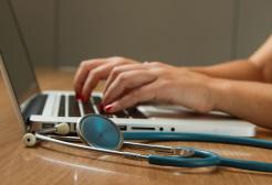 A woman typing on a laptop with a stethoscope next to her