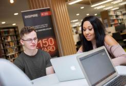 A couple of students looking at the screen of a MacBook together, inside the university’s Clifford Whitworth Library