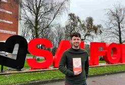 Pic shows Josh with book standing in front of the Salford sign