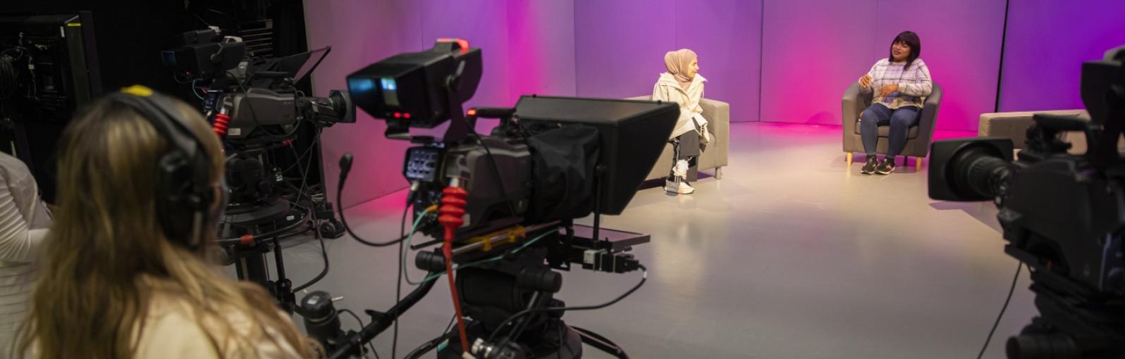Two female students presenting a chat show being filmed by another student 