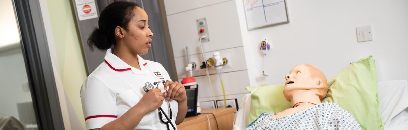 A female student nurse holding a stethoscope looking at a patient manikin