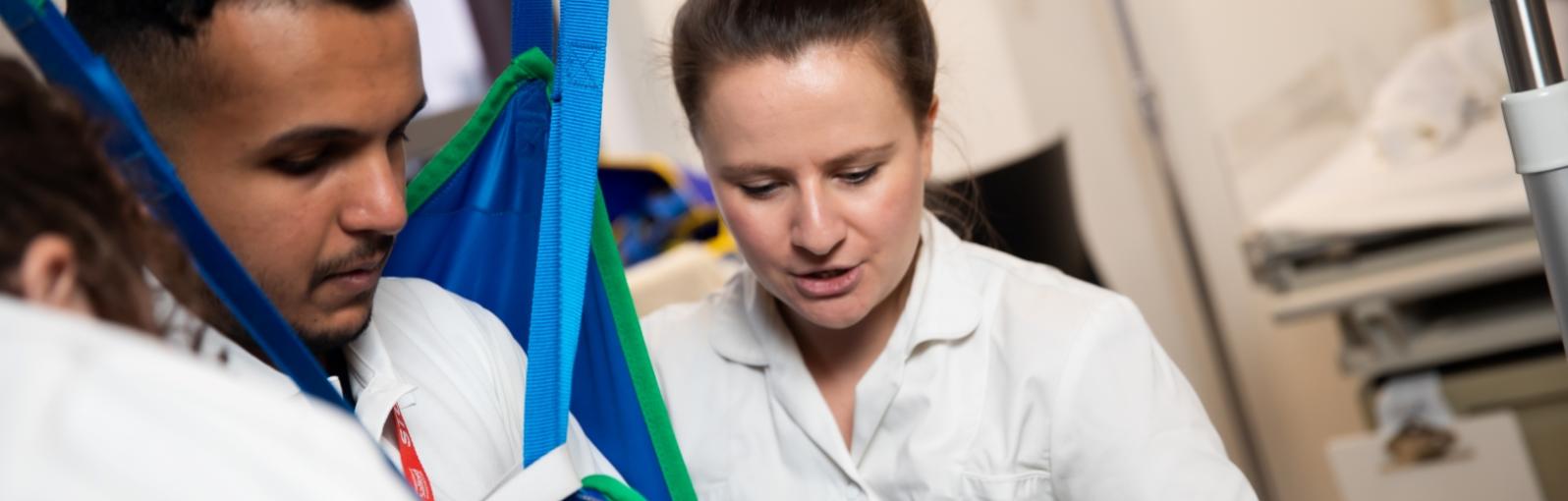 Male and female Occupational Therapy students practising moving and handling techniques with a sling and hoist under the direction of a tutor.