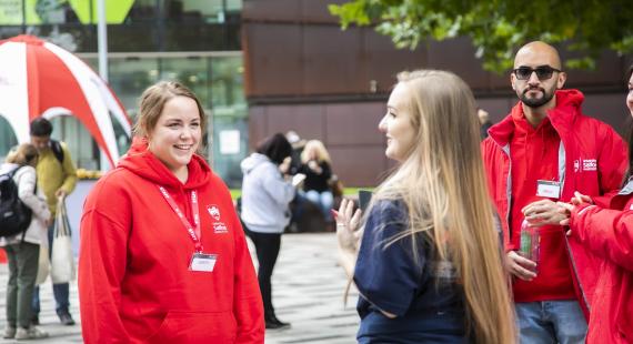 Three student ambassadors, in red University-branded hoodies, chatting to member of staff with concrete University building in the background