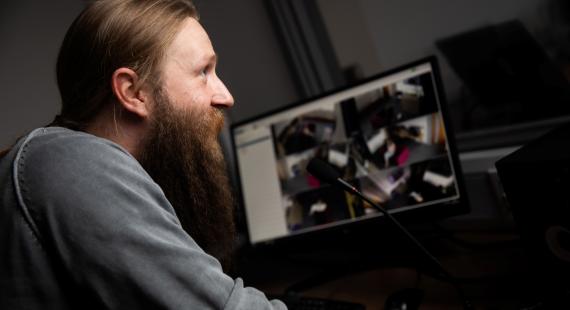  A man with a long beard and ponytail is speaking into a microphone while looking at a computer monitor displaying multiple surveillance camera feeds.