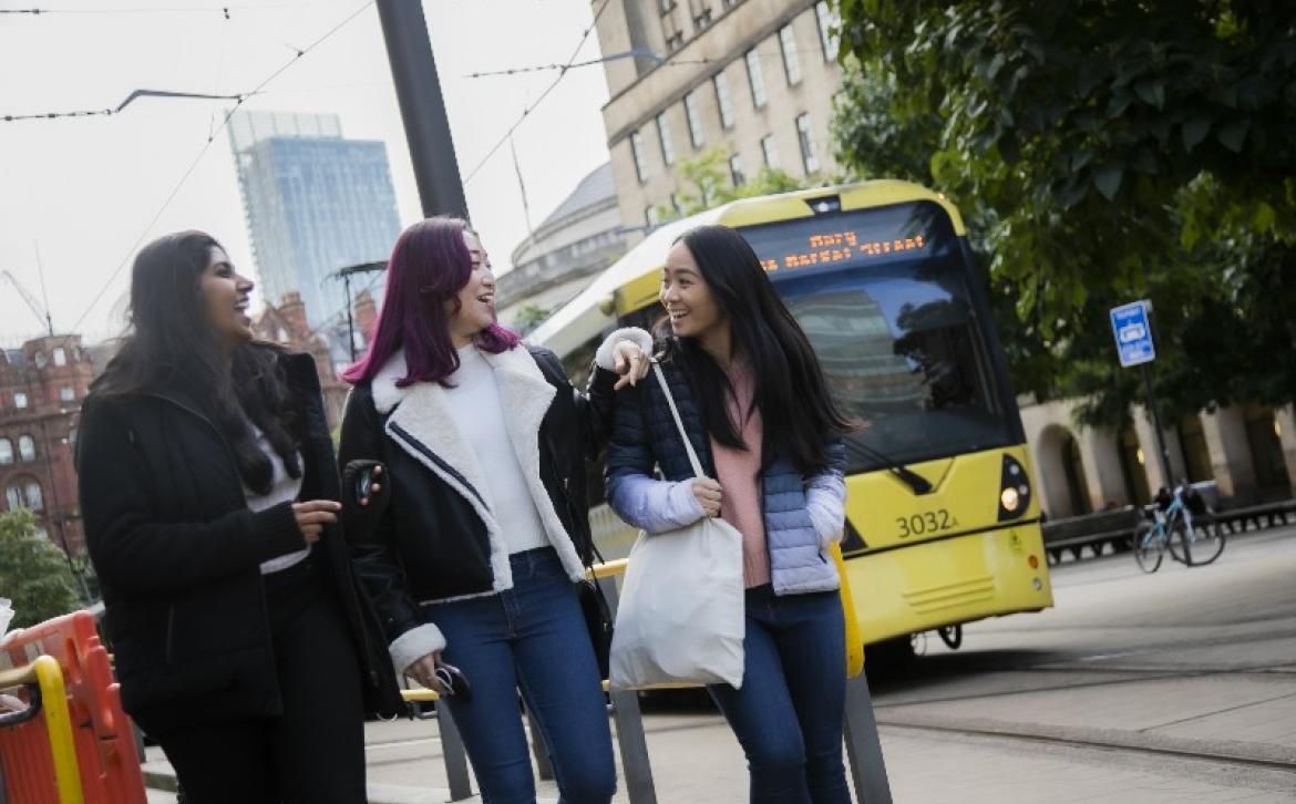 Three female students chatting as a yellow tram drives past behind them