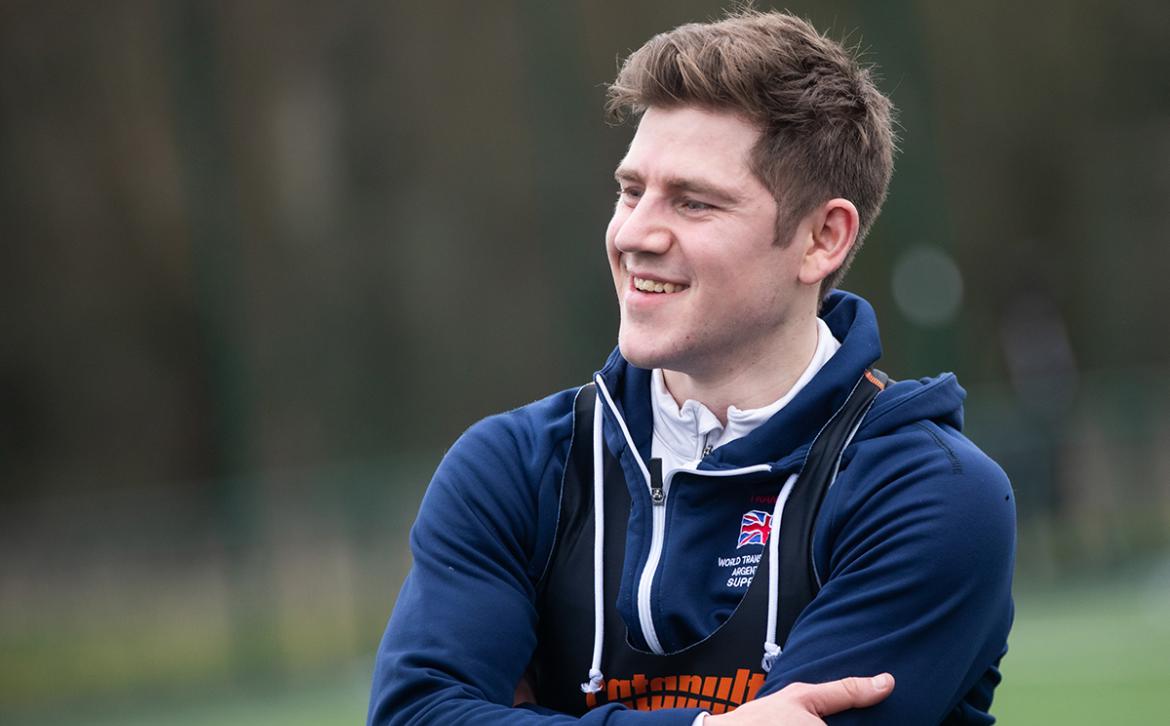 Sam wearing a navy hoodie and GPS tracking vest stood on the outdoor 3G pitch smiling to one side.