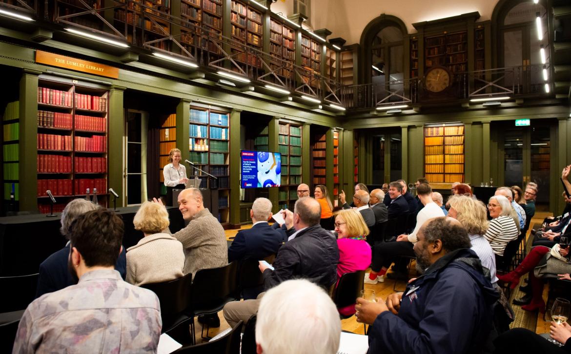 A presenter stands on a raised stage with an audience filling seats in a large library. The presenter is smiling at the audience who are looking at each other and smiling too